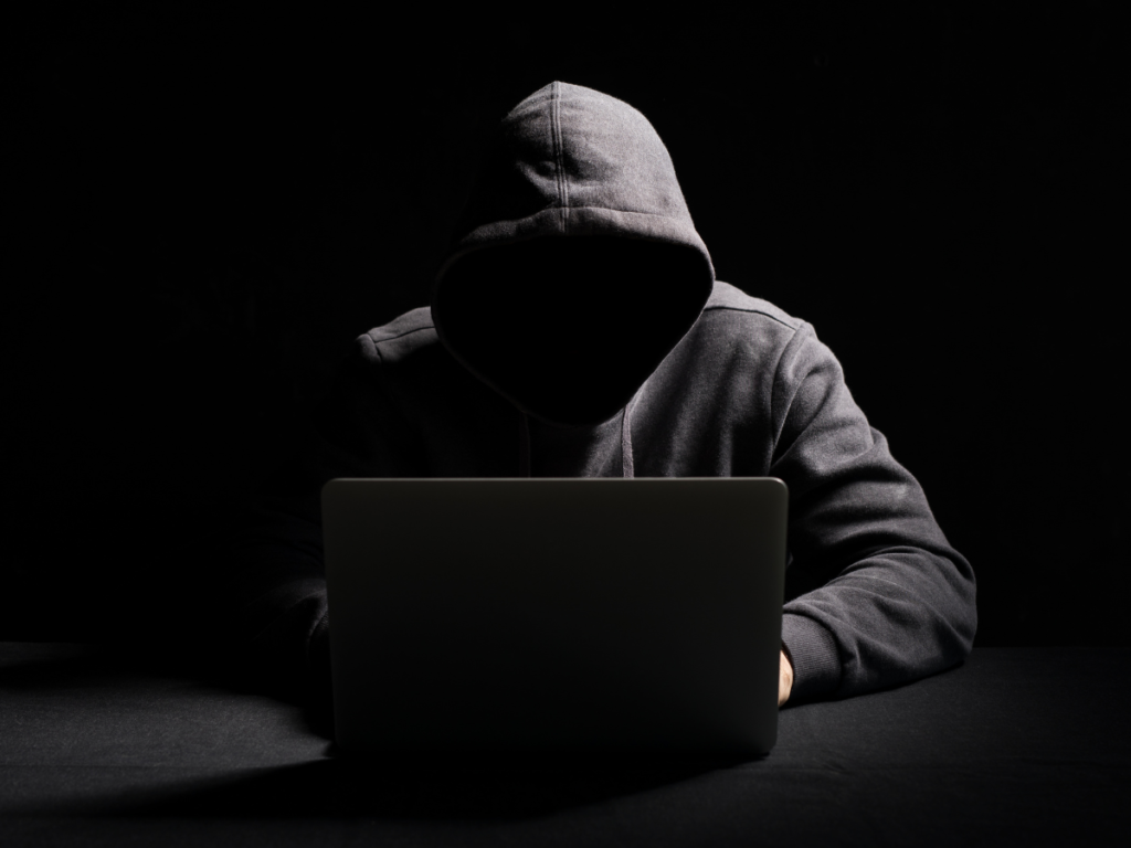 A man in a hoodie with his face hidden is using a laptop in a dark room.