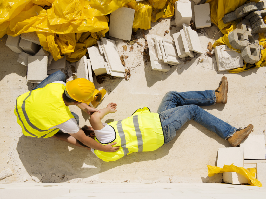 Construction worker on the ground of a construction site in pain being aided by another construction worker.
