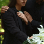 Mourners in black clothing standing beside white lilies at a funeral, with hands placed on their chest in grief.