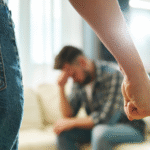 lose-up of a clenched fist in the foreground with a distressed man sitting on a couch in the background, symbolizing domestic violence and intimidation.