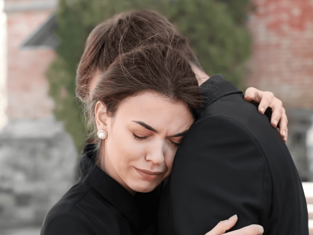 Grieving woman with closed eyes hugging a man in black clothing, showing comfort and sorrow.