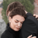 Grieving woman with closed eyes hugging a man in black clothing, showing comfort and sorrow.