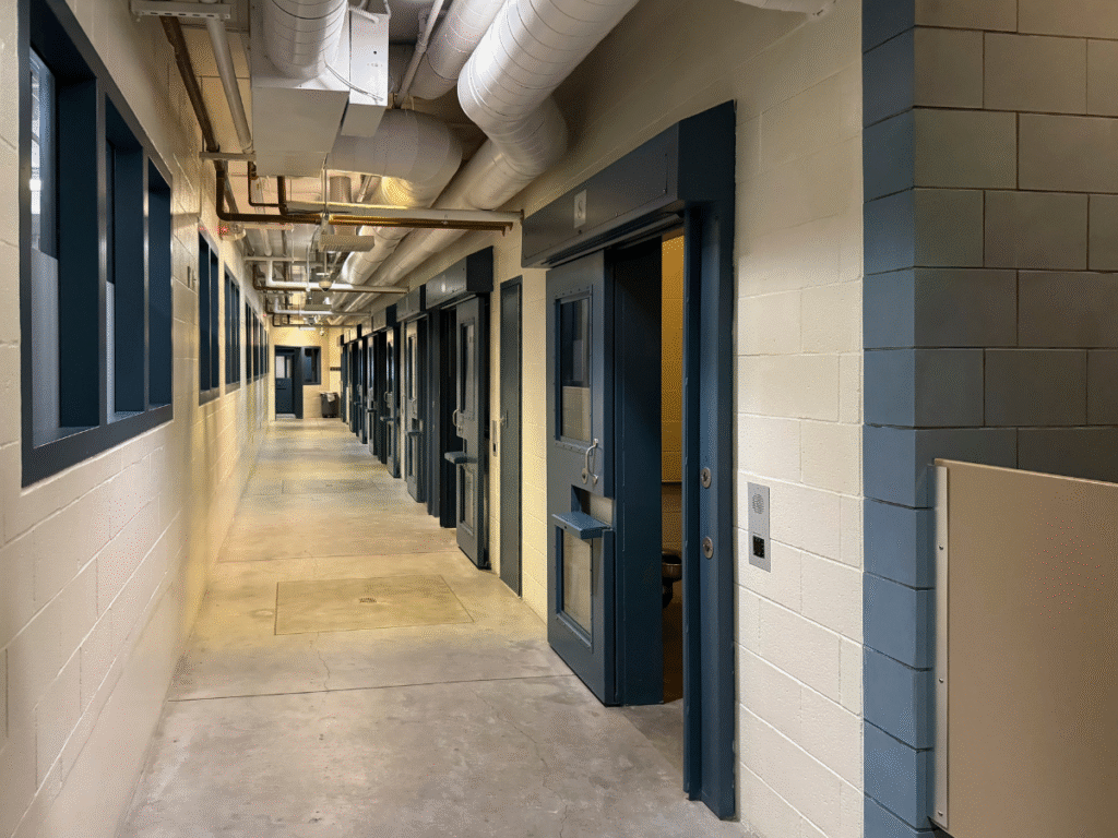 A narrow hallway lined with secured cell doors inside a Wyoming correctional facility, representing the difference between jail and prison environments.