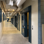 A narrow hallway lined with secured cell doors inside a Wyoming correctional facility, representing the difference between jail and prison environments.