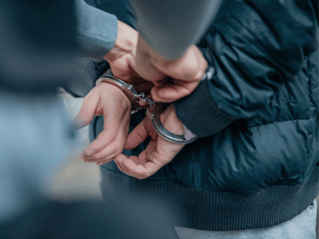 A person in a dark jacket is shown from behind with their hands cuffed together as a law enforcement officer secures the handcuffs, representing an arrest related to an out-of-state warrant.
