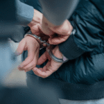 A person in a dark jacket is shown from behind with their hands cuffed together as a law enforcement officer secures the handcuffs, representing an arrest related to an out-of-state warrant.
