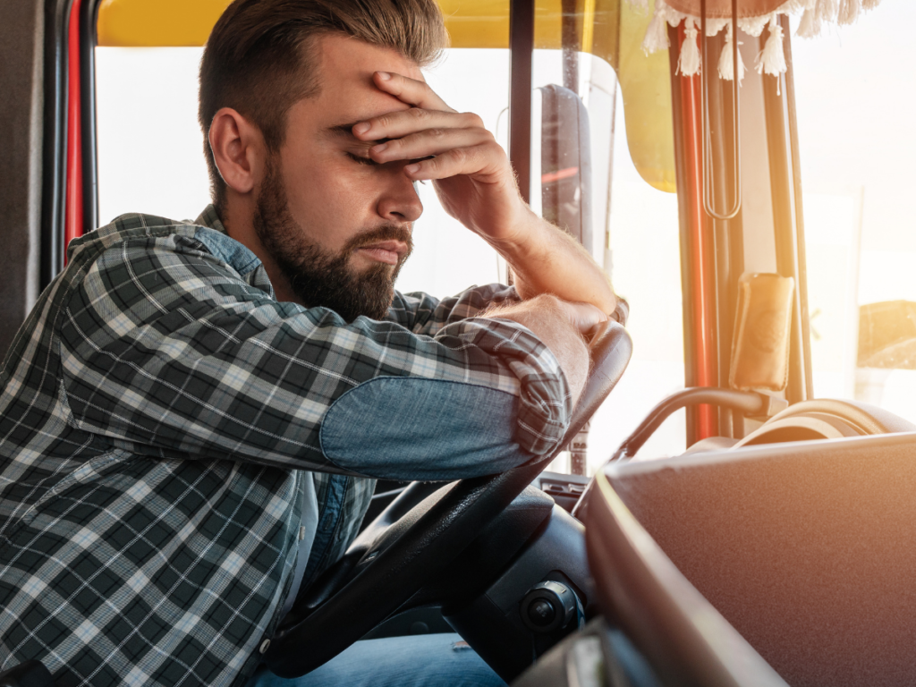 A fatigued truck driver sits behind the wheel with eyes closed and hand on his forehead, illustrating the dangers of drowsy driving and how exhaustion can contribute to serious trucking accidents.