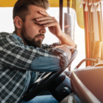 A fatigued truck driver sits behind the wheel with eyes closed and hand on his forehead, illustrating the dangers of drowsy driving and how exhaustion can contribute to serious trucking accidents.