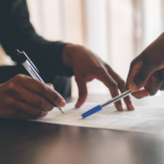 Person signing a document while another points to sections on the page, representing insurance paperwork and disputes over liability after an accident.