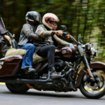 Two motorcyclists wearing helmets ride together on a touring motorcycle along a wooded road, suggesting travel and group riding.