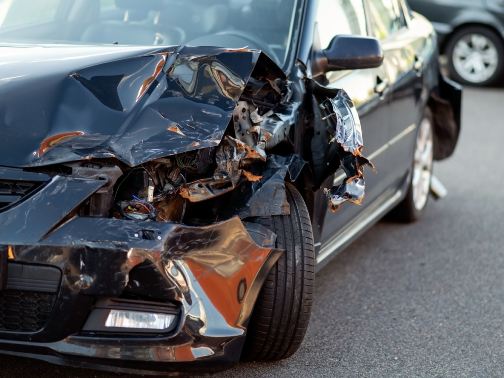 Front-end damage to a black passenger vehicle after a collision, showing a crushed front-left corner, broken headlight, exposed internal components, and significant body damage while parked at the accident scene.
