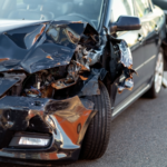 Front-end damage to a black passenger vehicle after a collision, showing a crushed front-left corner, broken headlight, exposed internal components, and significant body damage while parked at the accident scene.