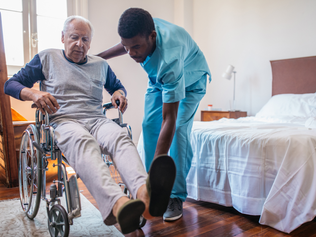 Caregiver assisting an elderly nursing home resident in a wheelchair beside a bed, illustrating a blog about nursing home responsibility for resident-on-resident abuse and neglect in Wyoming.