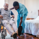 Caregiver assisting an elderly nursing home resident in a wheelchair beside a bed, illustrating a blog about nursing home responsibility for resident-on-resident abuse and neglect in Wyoming.